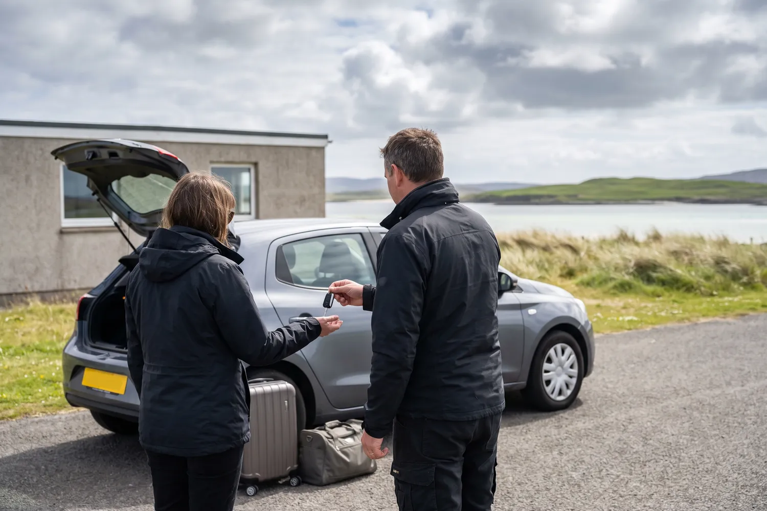 A traveller with a suitcase being handed car keys beside a hire car on the apron of a very small island airfield above a pale sandy bay