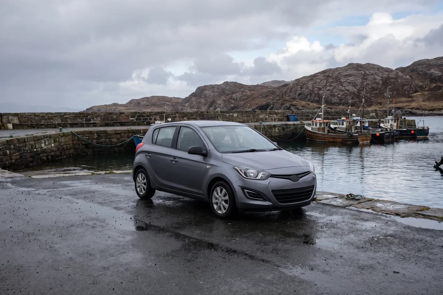 A single hire car parked alone on wet tarmac at a quiet Hebridean harbour, with moored fishing boats and bare rocky hills behind