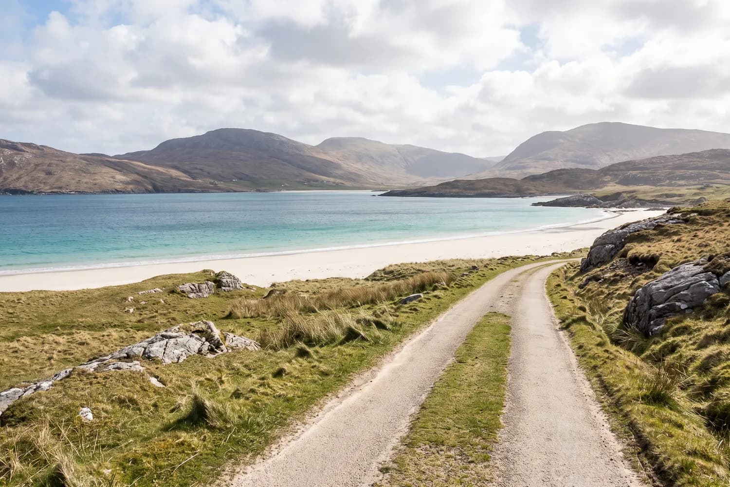 A single-track road curving above a wide white shell-sand bay with bare hills on the far shore