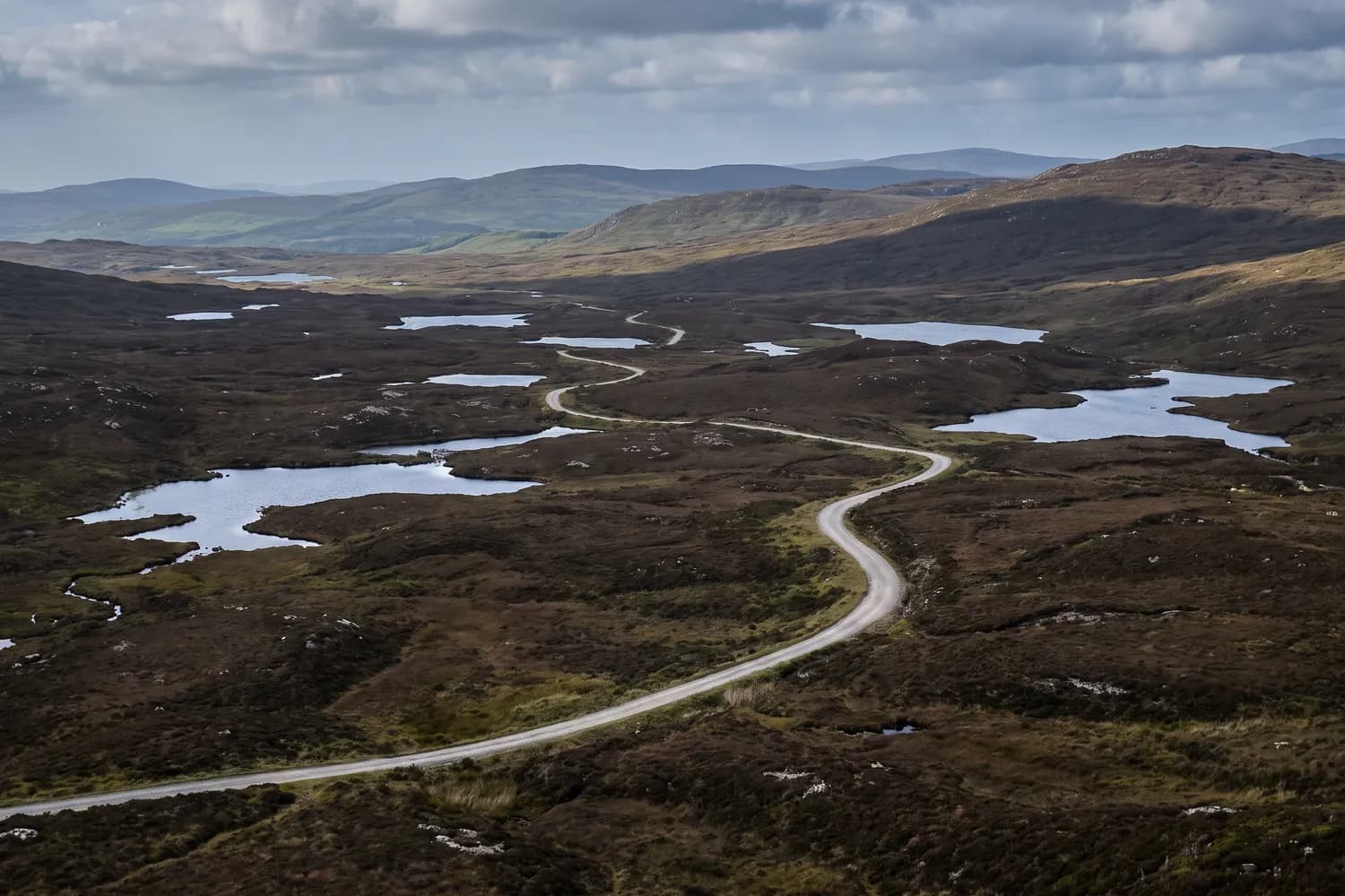 A single-track road threading between peat lochans across open moorland, with low ridges receding into haze