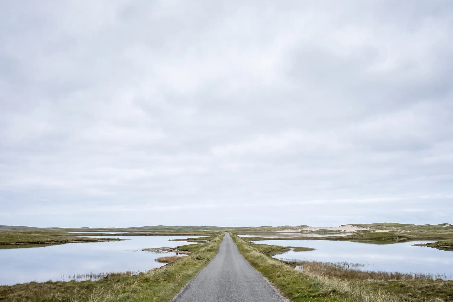 A low straight road crossing flat machair between shallow freshwater lochs that mirror the sky