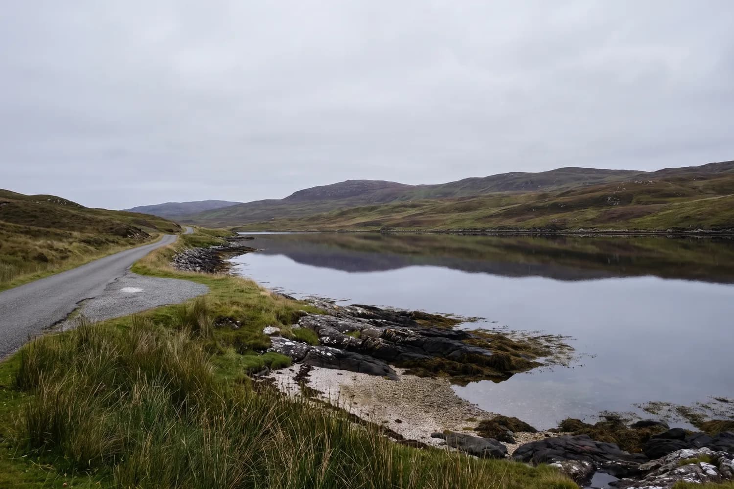 A gravel passing place beside a still sea loch holding the reflection of low hills on the far shore