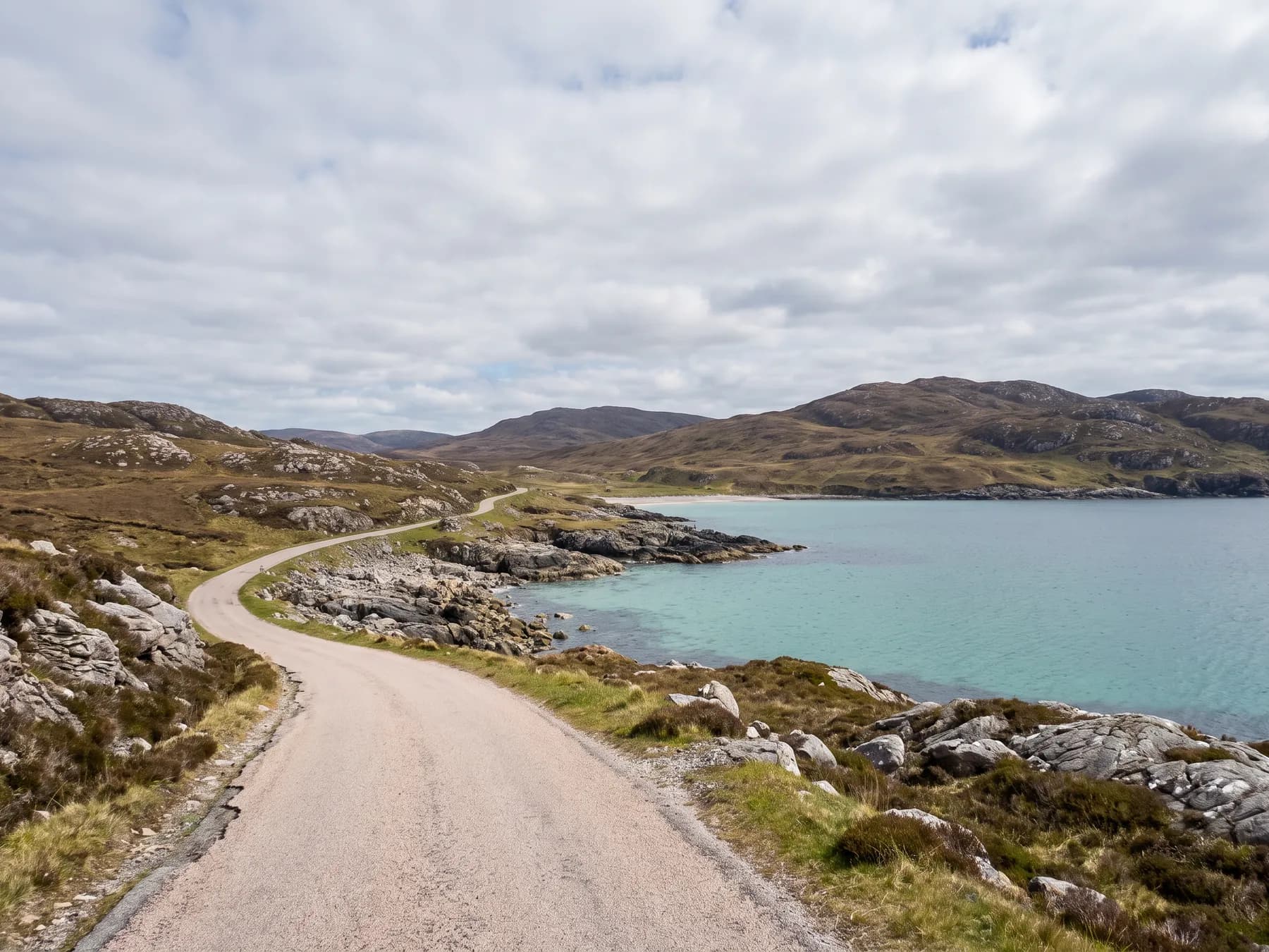 A single-track road curving along a rocky Hebridean shoreline above a shallow turquoise bay