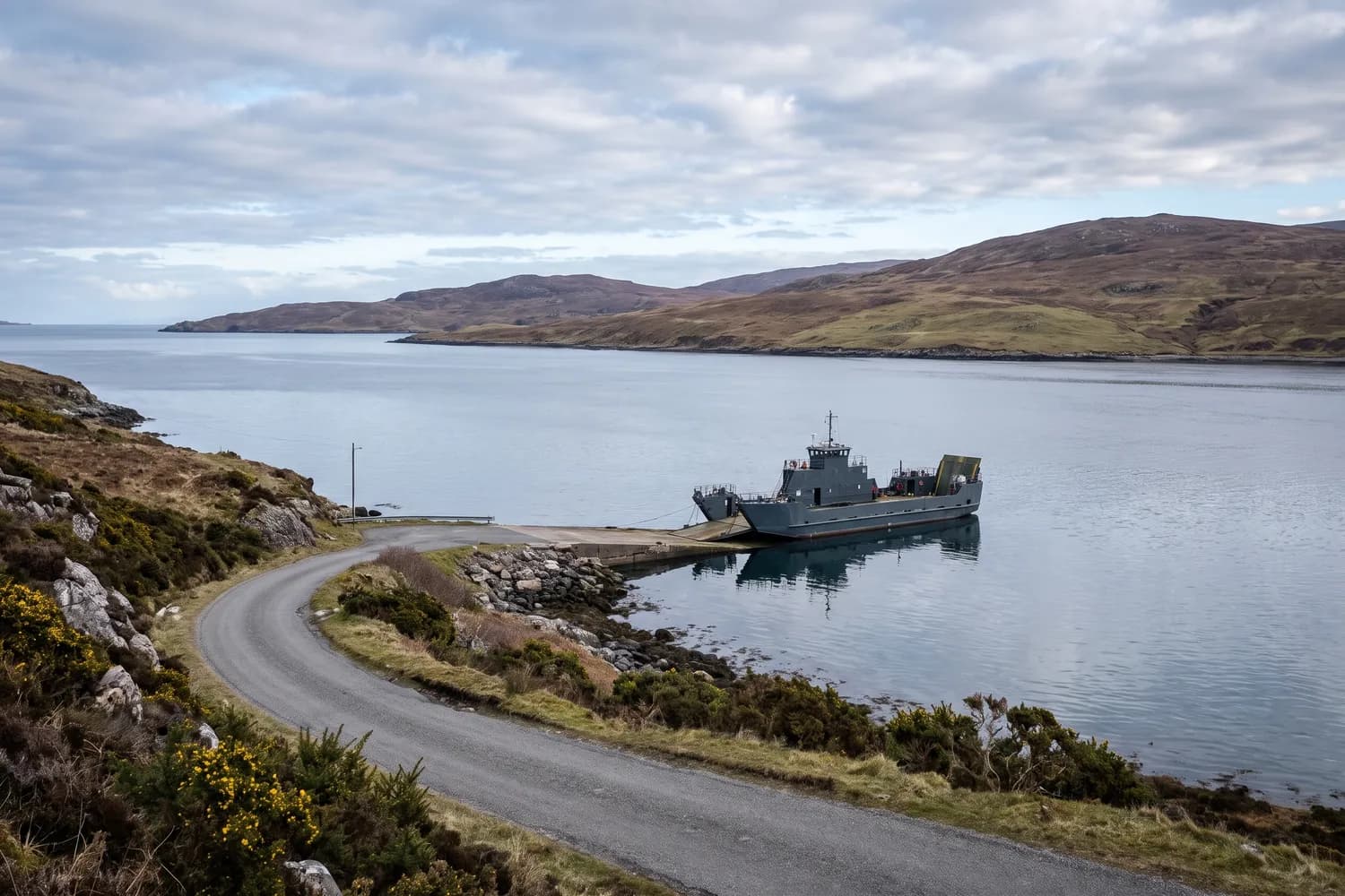 A coastal road descending towards a ferry slipway where a plain-hulled vessel lies at the linkspan