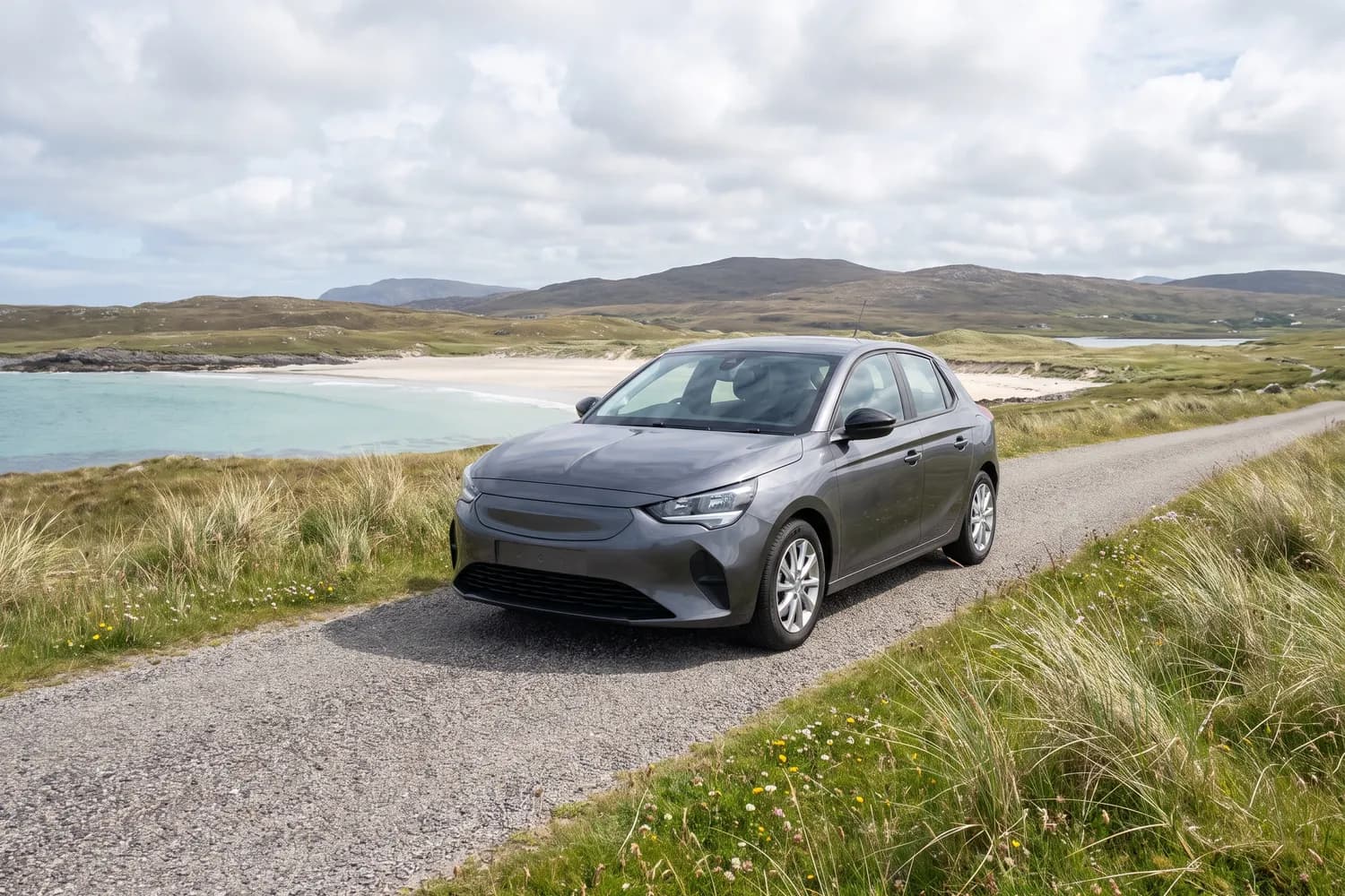 A small hatchback parked in a gravel passing place on a machair road above a pale sandy bay
